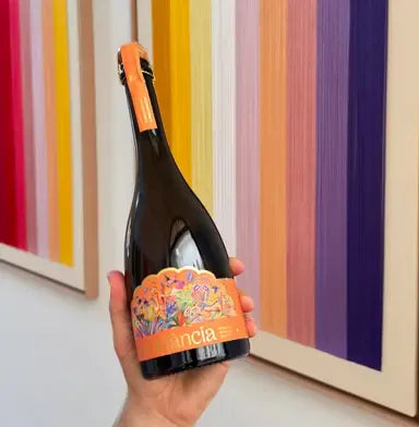 A hand holding a bottle of Delância orange botanical sparkling wine against a vibrant striped backdrop